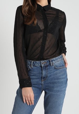 Femme portant un chemisier noir transparent à manches longues avec des pois texturés, boutonné sur un soutien-gorge noir, associé à un jean bleu taille haute.