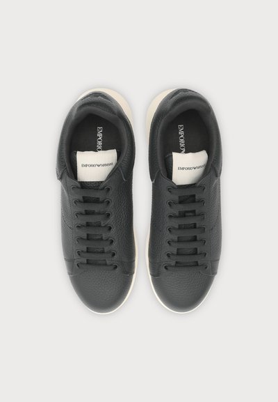 Paire de baskets en cuir noir à lacets avec semelles blanches et étiquettes blanches de marque sur la languette, vues de dessus sur un fond clair.
