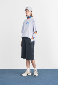 Chemise à manches courtes violette clair avec un graphique "23", shorts noirs et baskets beiges. Casquette grise ; accents multicolores sur les manches.