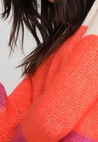 Strickpullover mit kräftigen Streifen in Orange, Pink und Creme. Strukturierter Stoff mit lockerer Passform, der unterschiedlich breite Farbbereiche zeigt.