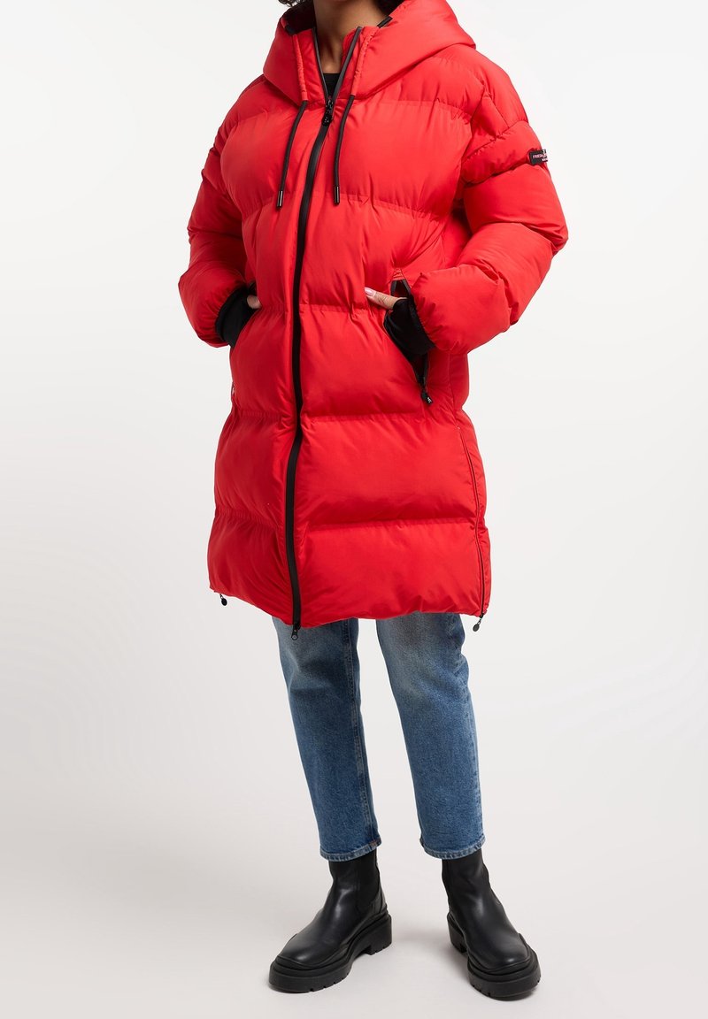 Giacca trapuntata rossa con cappuccio, chiusura a zip frontale e due tasche laterali. Presenta dettagli neri e una texture trapuntata. Abbinata a jeans blu e stivali neri.