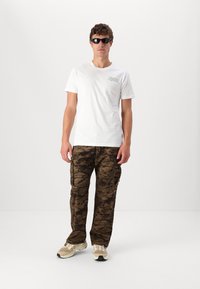 Hvid kortærmet t-shirt med grønt grafik, brune camouflage cargo bukser og beige sneakers. Solbriller tilføjer en markant accessory.