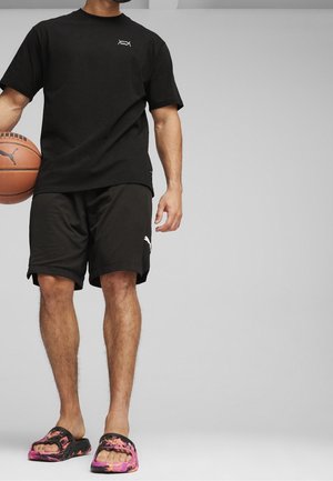 Mężczyzna ubrany w czarne spodenki Puma i czarną koszulkę trzymający koszykówkę, stojący boso w czarno-różowych klapkach.