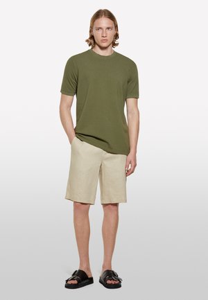 Olivgrünes, tailliertes T-Shirt, beiges Leinen-Shorts, schwarze flache Sandalen mit Schnallendetail, Modell steht mit Händen in den Taschen.