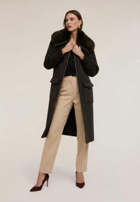 Lång mörk kappa med en fuskpälskrage, framdragkedja och två stora framfickor, i kombination med beige byxor och burgundy stilettklackar.