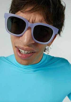 Lunettes de soleil violettes surdimensionnées avec des verres foncés, portées avec un haut de maillot de bain turquoise ajusté, affichant une texture lisse et un design moderne.