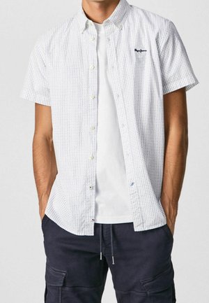 Mies pukeutuneena valkoiseen, lyhythihaiseen nappipaitaan, jossa on pieniä sinisiä pisteitä, valkoinen T-paita alla ja tummat cargo-housut, kädet taskuissa.