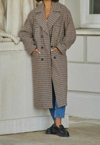 Lange geruite jas in bruine en zwarte tinten, met een dubbelgeknoopt ontwerp, oversized revers en zijzakken, gecombineerd met donkere schoenen en blauwe jeans.