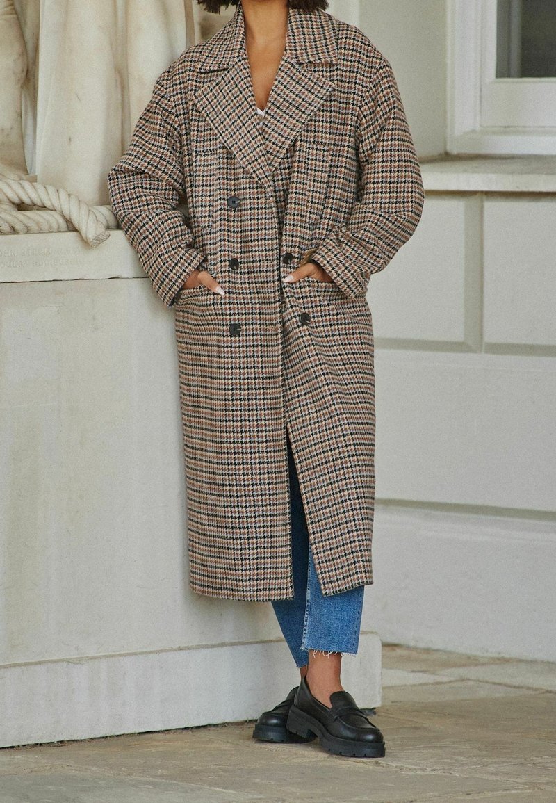 Lange geruite jas in bruine en zwarte tinten, met een dubbelgeknoopt ontwerp, oversized revers en zijzakken, gecombineerd met donkere schoenen en blauwe jeans.