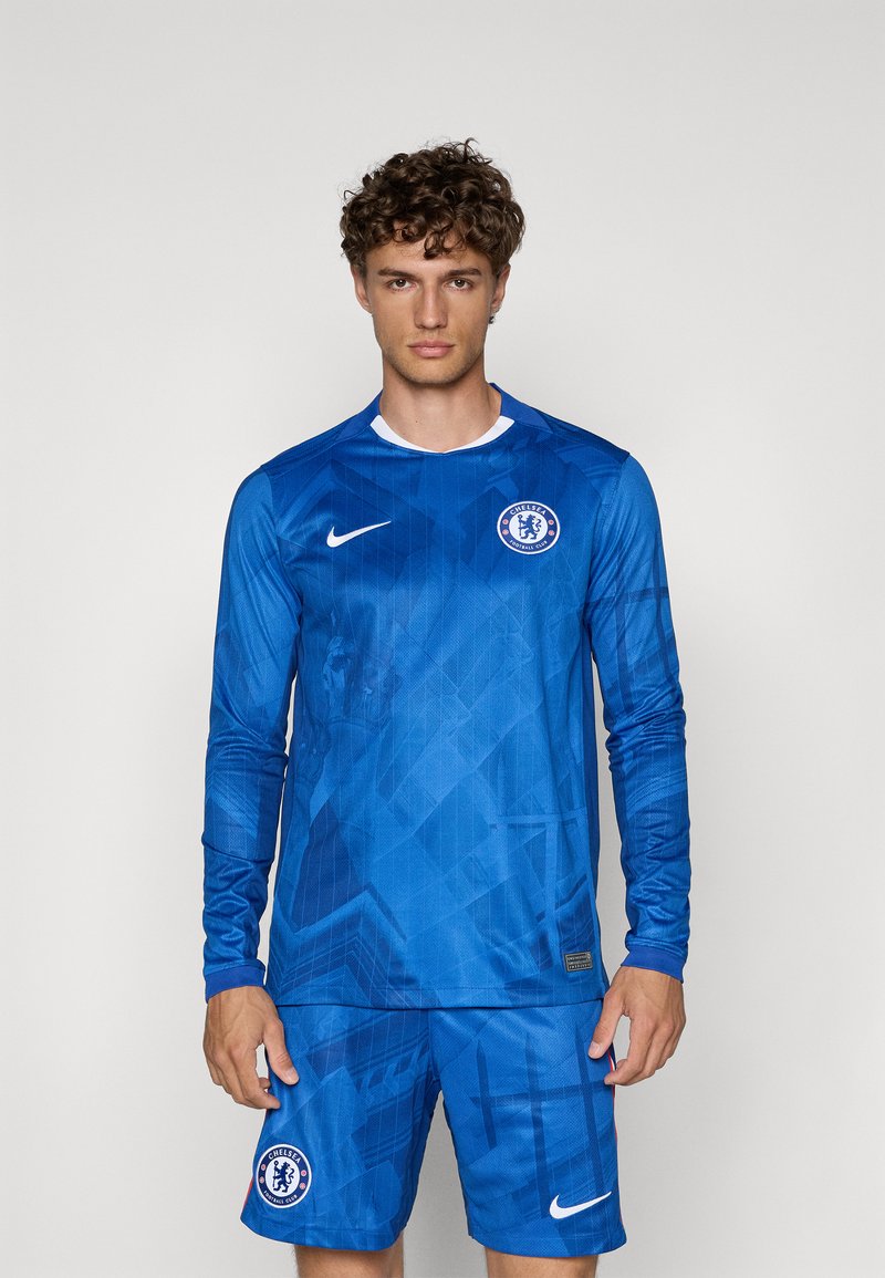 Blåt fodboldoutfit med langærmet trøje og shorts. Tekstureret stof med geometriske mønstre, hvidt Nike-logo og Chelsea FC-emblem.