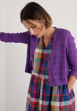 Cardigan en maille violette avec un motif texturé, associé à une robe à carreaux colorée avec un décolleté en V et une taille froncée.