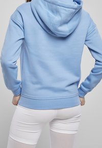 Ljusblå hoodie i mjukt tyg, med huva, ribbade ärmslut och midja. Designen är enkel utan synliga mönster.