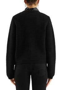 Person, der einen schwarzen strukturierten Strickpullover und schwarze Hose trägt, mit dem Gesicht abgewandt, kurzen Haaren und sichtbaren Kettenohrringen.