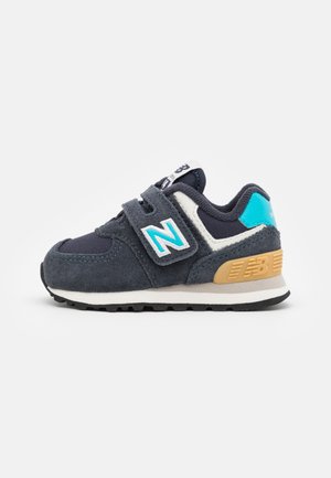 Marinblå ruskinds sneakers med velcro-lukning, hvide og turkise accenter, lys tan hæl og sort gummisål. Retro design med "N" logo.