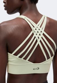 Gros plan du dos d’une personne portant un soutien-gorge de sport clair avec plusieurs bretelles croisées et un petit logo infini sur la bande.