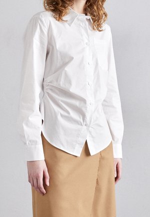 Kiharatukkainen nainen, joka käyttää valkoista napillista paitaa ja vaaleanruskeaa hametta, jossa on edessä raakareunainen sauma, seisoo yksinkertaisen vaaleanharmaan taustan edessä.