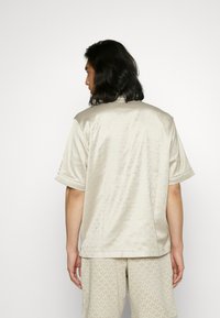 Ljust beige kortärmad skjorta i glänsande satin med ett subtilt bladmönster, med en rundad krage och uppvikt ärmar.