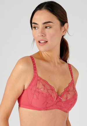 Woman with brown hair in a low ponytail wearing a coral pink lace bra with floral embroidery, looking slightly to the left against a plain background.