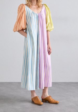 Kobieta w długiej, wielokolorowej sukience w paski z bufkami na rękawach w kolorach pomarańczowym i żółtym, nosząca beżowe sandały typu mule z wiązaniami.