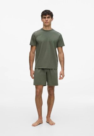 Giovane uomo in piedi a piedi nudi che indossa una camicia a maniche corte e pantaloncini verde oliva coordinati su uno sfondo bianco semplice.