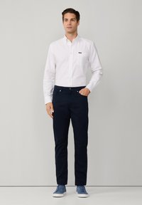 Camicia bianca con bottoni e taschino sul petto, abbinata a pantaloni slim-fit blu scuro. L'uomo indossa scarpe da ginnastica blu e si trova davanti a uno sfondo grigio neutro.