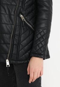 Chaqueta de cuero negra acolchada con costuras diagonales, cremallera plateada y un bolsillo con cremallera en ángulo en el lateral.