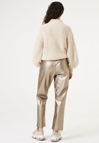Chunky gebreide crème trui met een hoge kraag en kabelpatronen, gecombineerd met glanzende gouden broek en lichte sneakers.