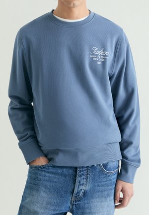 Hombre con sudadera azul de cuello redondo con texto blanco "Scalpers Sunset & Design New Code 1990" y vaqueros azules, con una mano en el bolsillo.