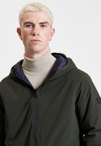 Giacca a vento verde scuro con una texture liscia, chiusura con zip e fodera blu navy, indossata sopra un dolcevita beige chiaro.