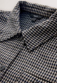 Tessuto di camicia a quadri grigio e blu navy con un aspetto strutturato; presenta un colletto a punta e un bottone nero. Etichetta del marchio visibile all'interno.