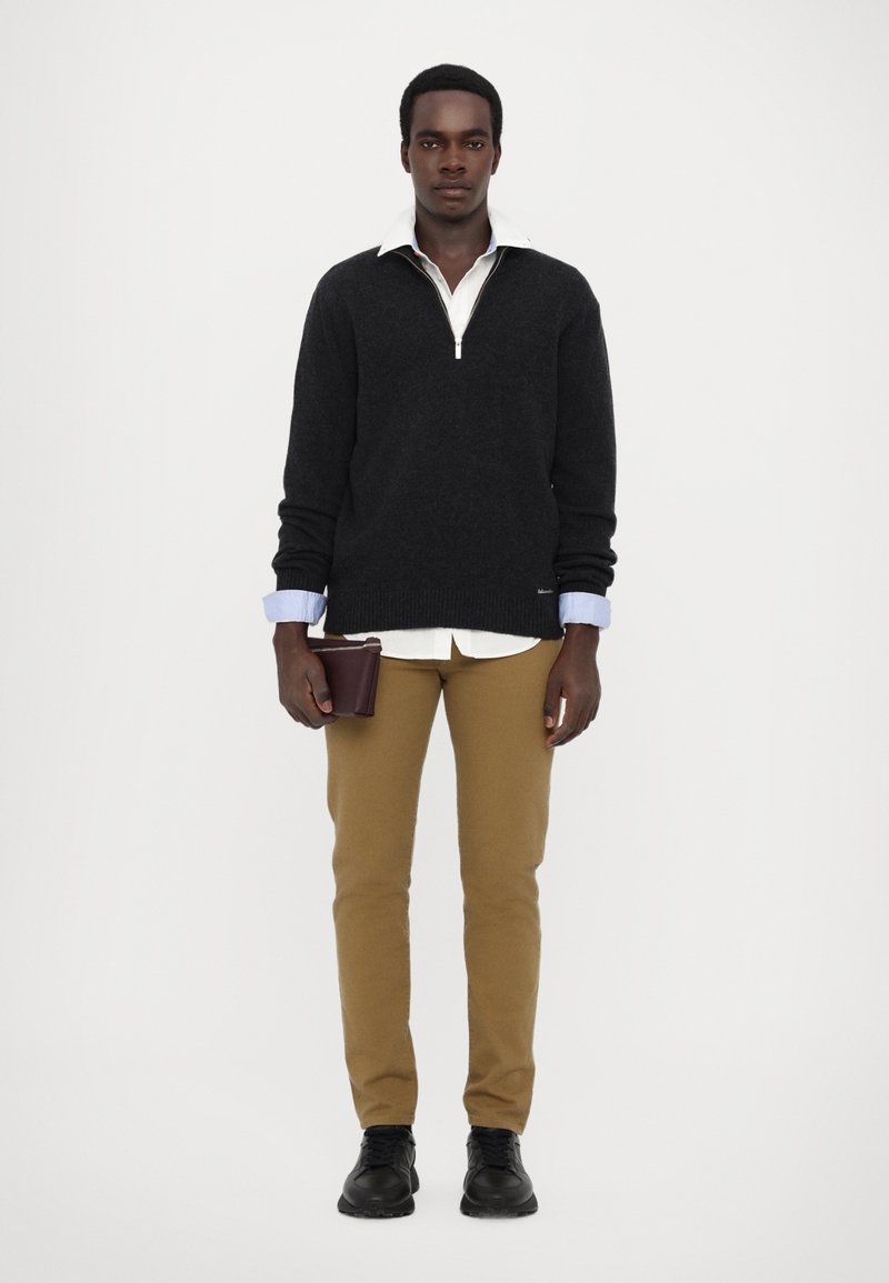 Svart zip-up sweatshirt, krämfärgad skjorta med krage under, beiga byxor, håller en mörk plånbok, svarta skor, slätt tyg, avslappnad passform.