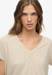 Beige V-Ausschnitt T-Shirt aus weichem Stoff, mit kurzen Ärmeln und dezentem Logo-Stick auf der Brust. Glatte Textur mit lockerer Passform.