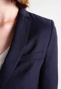 Blazer bleu marine en tissu texturé, coupe à simple boutonnage avec un détail de poche discret. Coupe ajustée pour un look cintré.