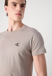 Smėlinis medvilninis marškinėliai su apvalia iškirpte ir trumpomis rankovėmis; kairėje krūtinės pusėje yra juodas siuvinėtas "cK" logotipas.