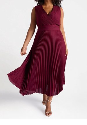 Kobieta ubrana w bezrękawnikową, głęboko czerwoną plisowaną sukienkę midi z dekoltem w szpic i dopasowaną talią, spacerująca w beżowych sandałach na paski.