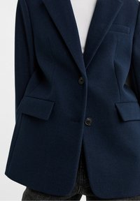 Marineblauer Wollblazer mit strukturiertem Design, mit Schlitzrevers, zwei aufgesetzten Taschen vorne und schwarzen Knöpfen. Der Stoff hat ein weiches, texturiertes Aussehen.