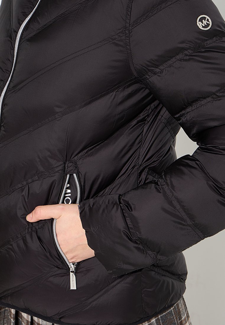 Veste matelassée noire avec des coutures diagonales, poches zippées et un logo argenté sur la manche gauche. Tissu lisse et brillant.