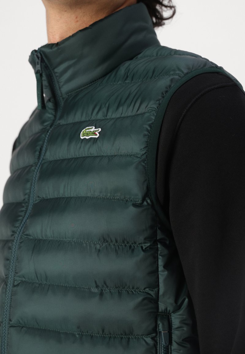 Zielona, pikowana kamizelka z zamkami błyskawicznymi, wysokim kołnierzem i małym haftowanym logo. Materiał wydaje się błyszczący i gładki. Pod spodem czarna koszula z długim rękawem.