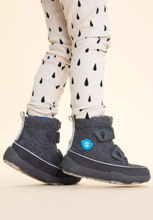 Child wearing grey winter boots with Velcro straps and white leggings patterned with black teardrops, standing on beige floor.