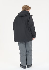 Zwarte waterdichte jas met een capuchon, gecombineerd met grijze broek. De jas heeft een losse pasvorm en beschikt over een rugpand.