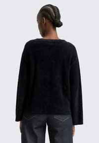 Schwarzer, flauschiger Pullover mit langen Ärmeln und weitem Ausschnitt, kombiniert mit dunklen Jeans. Die Textur wirkt weich und gemütlich.