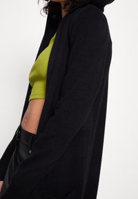 Czarny kardigan z kapturem o miękkiej fakturze, z otwartym przodem i dwoma bocznymi kieszeniami, zestawiony z zielonym prążkowanym crop topem.