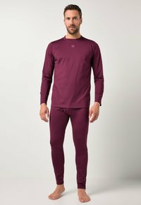 Uomo che indossa un top aderente a maniche lunghe e leggings di un viola scuro, in piedi scalzo su uno sfondo chiaro e semplice.