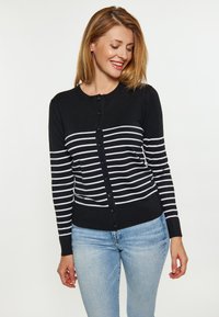 Cardigan nero con strisce orizzontali bianche, presenta un collo rotondo, chiusura con bottoni e polsini a costine, abbinato a jeans blu chiaro.