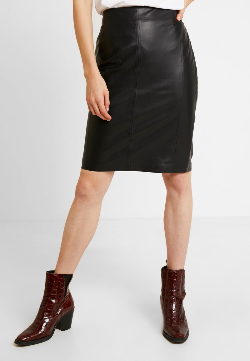 Jupe crayon en cuir noir avec des coutures apparentes, associée à des bottines à motif crocodile marron foncé et un haut blanc uni.