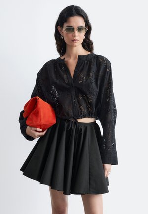Femme portant une blouse noire transparente boutonnée et une jupe noire tenant une pochette rouge vif pliée, avec des lunettes de soleil et des cheveux ondulés foncés.