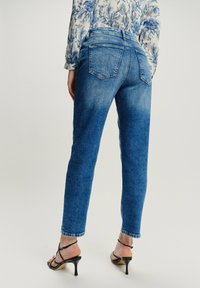 Kobieta w niebieskich dżinsach o średnim stanie i czarnych sandałach na obcasie, w zestawieniu z białą bluzką w niebieskie kwiatowe wzory.