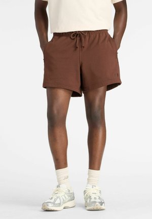Person trägt braune Shorts mit Kordelzug, weiße Kniestrümpfe und weiße und silberne Sportschuhe, steht mit den Händen in den Taschen.