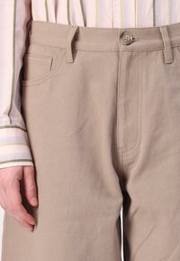 Beige bukser i en teksturert bomullsstoff, med glidelås foran, belte loops og sidelommer. Ved siden av en lysestripet skjorte.