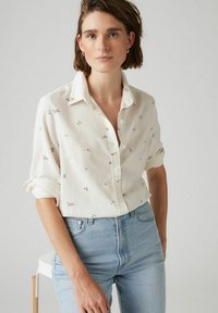 Camicia bianca con bottoni, caratterizzata da ricami floreali in miniatura, maniche arrotolate e vestibilità rilassata, abbinata a jeans high-waisted di colore azzurro chiaro.